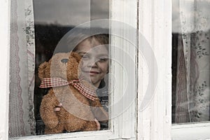 Little grimy boy looking through a dirty old window. A sad smile on his face. He is shows a brown bear