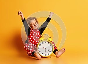 Little happy blond girl in red dotted pajamas sitting on floor with big alarm clock and feeling excited with raised hands