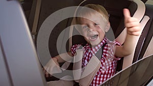 Little happy boy is sitting in a child car seat in a car and shows his thumb up.