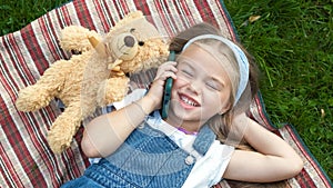 Little happy child girl laying on a blanket on green lawn in summer with her teddy bear talking on mobile phone