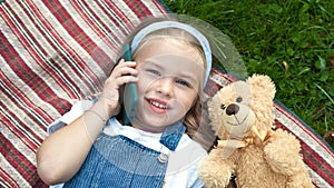 Little happy child girl laying on a blanket on green lawn in summer with her teddy bear talking on mobile phone