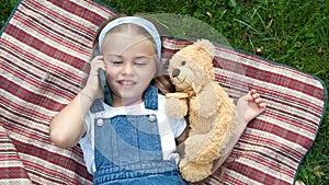 Little happy child girl laying on a blanket on green lawn in summer with her teddy bear talking on mobile phone