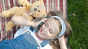 Little happy child girl laying on a blanket on green lawn in summer with her teddy bear talking on mobile phone