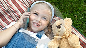 Little happy child girl laying on green lawn with her teddy bear talking on mobile phone