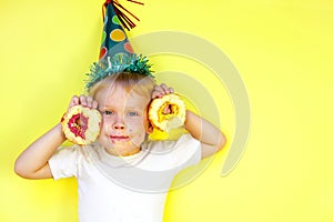 Little happy cute boy in birthday hat smiling and holding two bitten donuts near his face. Cheerful child playing with donuts on y