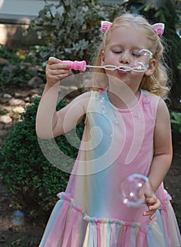 Little happy funny girl with blond hair blowing soap bubbles in amusement park