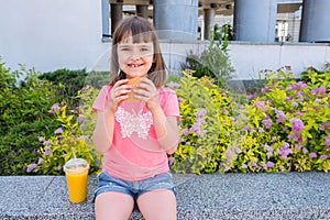 The little happy girl with cheeseburger in hands looks in the camera. Fast fat food