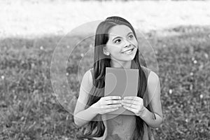 Little happy girl reading book outdoors sunny day, daydreamer concept