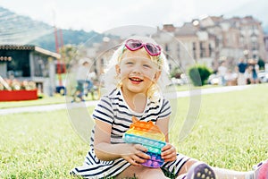 Little happy girl in sunglasses sitting on the lawn with anti-stress pop it smiles.