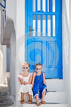 Little happy girls in dresses at street of typical greek traditional village on Mykonos Island, in Greece