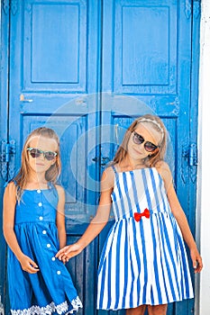 Little happy girls in dresses at street of typical greek traditional village on Mykonos Island, in Greece
