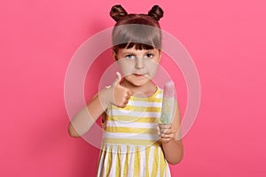 Little happy kid girl holding ice cream and showing thumb up while looking directly at camera, wearing summer dress, has two knots