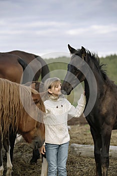 Little happy young girl standing among horses and foals in a white sweater jeans. Lifestyle portrait