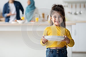 Little Helper. Cute Arab Girl Holding Plates In Kitchen, Smiling At Camera