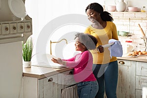 Little Helper. Cute Black Preschool Girl Washing Dishes With Mom In Kitchen