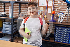 Little hispanic boy wearing student backpack and holding book at school class pointing thumb up to the side smiling happy with