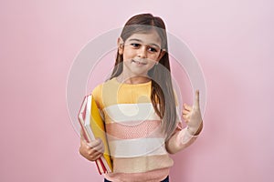 Little hispanic girl holding books smiling with an idea or question pointing finger with happy face, number one