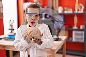 Little hispanic girl holding brain at science class at school afraid and shocked with surprise and amazed expression, fear and