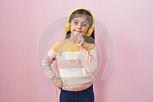 Little hispanic girl listening to music using headphones thinking concentrated about doubt with finger on chin and looking up