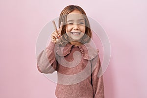 Little hispanic girl standing over pink background smiling with happy face winking at the camera doing victory sign