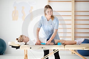 Little injure girl lying on massage table and exercising with young female doctor