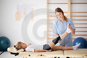 Little injure girl lying on massage table and exercising with young female doctor
