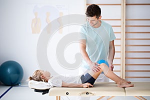 Little injure girl on massage table exercising with young male doctor