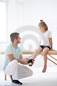 Little injure girl on massage table exercising with young male doctor