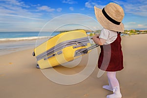 A little joyful cute girl in a blue summer dress and a straw hat holds a yellow suitcase. Vacation, holidays, trip to the sea,