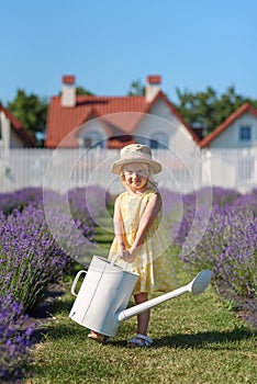 Little joyful girl in yellow dress with watering can at lavender garden. Smiling cute baby girl, summer joy, childhood