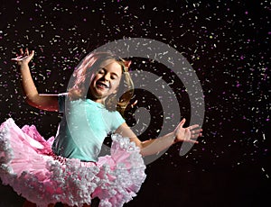Little kid in blue t-shirt and poofy skirt. Smiling, closed eyes, jumping up against black background, pink backlight. Close up