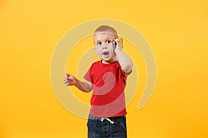 Little kid boy 3-4 years old in red t-shirt talking on mobile phone, conducting pleasant conversation isolated on yellow