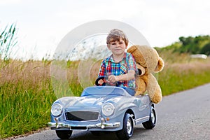 Little kid boy driving big toy car with a bear, outdoors.