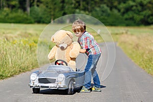 Little kid boy driving big toy car with a bear, outdoors.