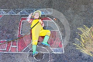 Little kid boy in fireman uniform having fun with fire truck picture drawing with colorful chalk on asphalt. From above