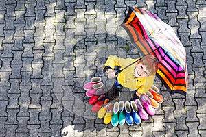 Little kid boy and group of colorful rain boots. Blond child standing under umbrella.