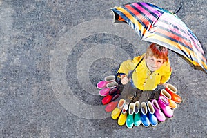 Little kid boy and group of colorful rain boots. Blond child standing under umbrella. Close-up of schoolkid and