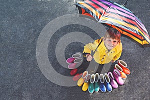 Little kid boy and group of colorful rain boots. Blond child standing under umbrella. Close-up of schoolkid and