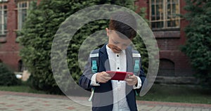 Little kid boy playing online game on smartphone while walking at street. Child in uniform with backpack lokking at