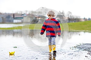 Little kid boy playing with paper boat by puddle