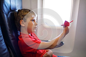 Little kid boy playing with red paper plane during flight on airplane. Child sitting inside aircraft by a window. Family