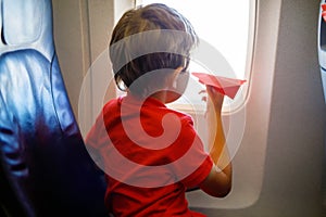 Little kid boy playing with red paper plane during flight on airplane