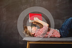 Little kid boy is reading a book while lying on bench on blackboard background. High speed of reading. Red book in hands