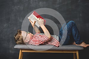 Little kid boy is reading a book while lying on bench on blackboard background. High speed of reading. Red book in hands