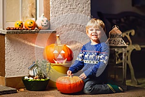 Little kid boy sitting with traditional jack-o-lanterns pumpkins for halloween by the decorated scary door, outdoors