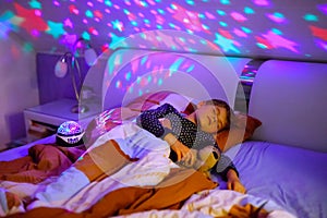 Little kid boy sleeping in bed with colorful lamp. School child dreaming and holding plush toy. Kid angry of darkness