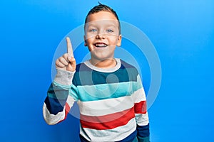 Little kid boy wearing striped sweater smiling with an idea or question pointing finger up with happy face, number one