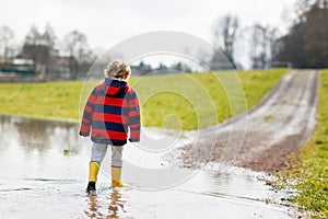 Little kid boy wearing yellow rain boots and walking and jumping into puddle on warm sunny spring day.