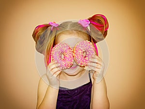 A little kid girl dabbles and plays with two fresh donuts before eating. A child holds donuts near his eyes and looks through the