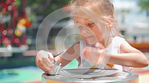 Little kid girl drink water jet of drinking fountain with defocused children on playground at background. Sunny summer